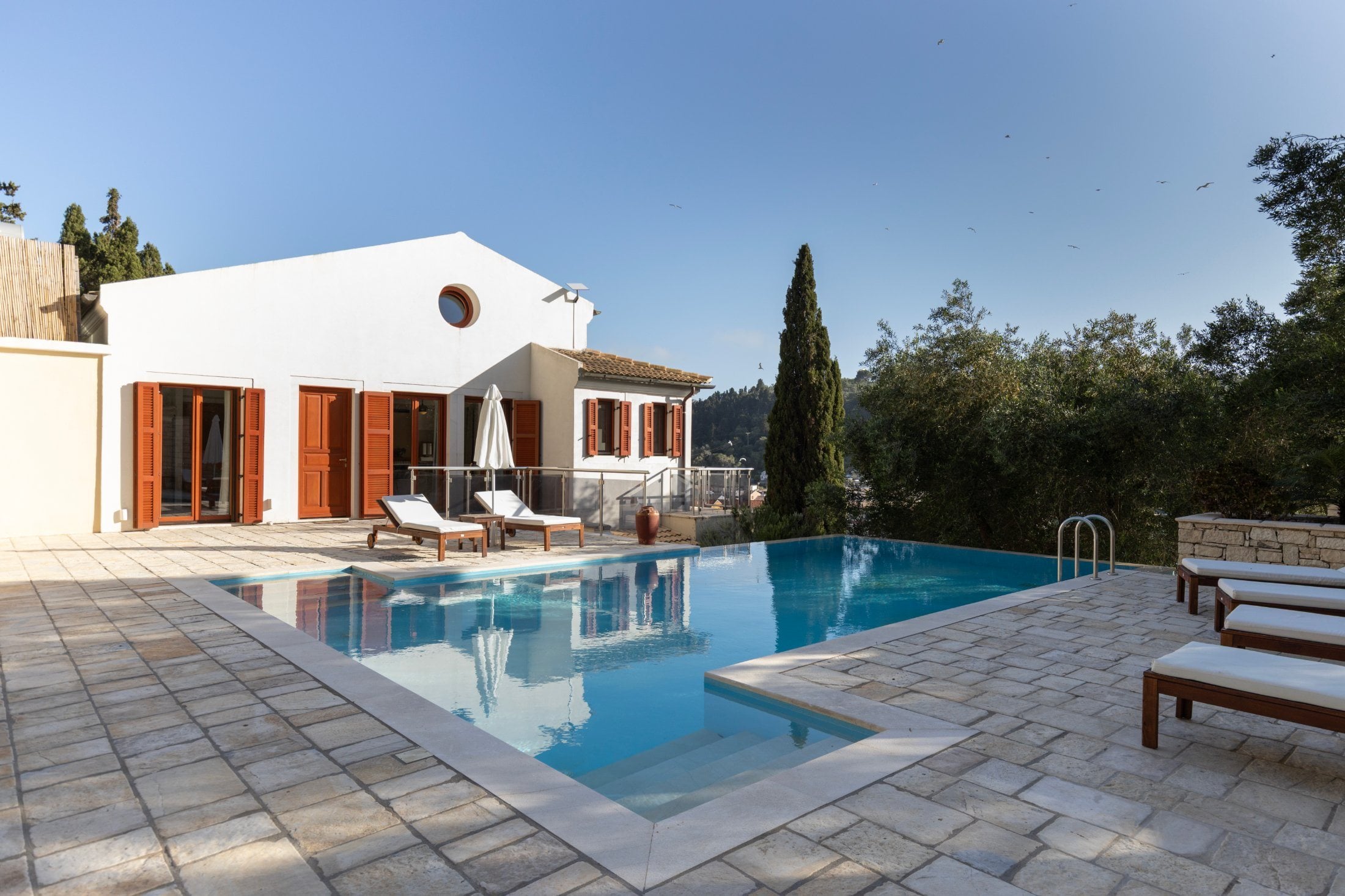 Infinity pool and stone terrace in front of the white villa with red shutters and cypress trees